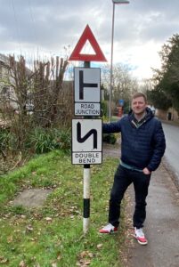 A photo of a man standing on the pavement next to a post with a vintage road sign on it.