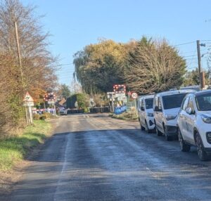 A road with cars and vans badly parked along it. The road is blocked by a closed level crossing in the distance.